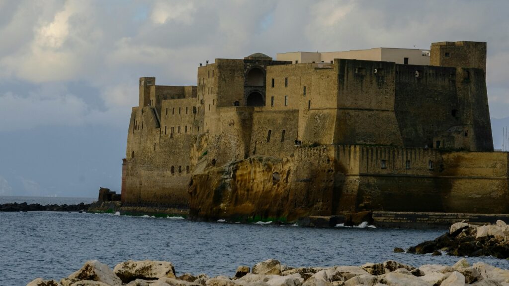 Scenic view of Castel dell'Ovo in Naples, Italy, with rocky shore and clouds.